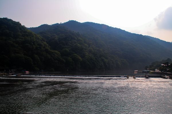 a wide river at the foot of a heavily forested green mountain, and a damn going across the river, thumbnail